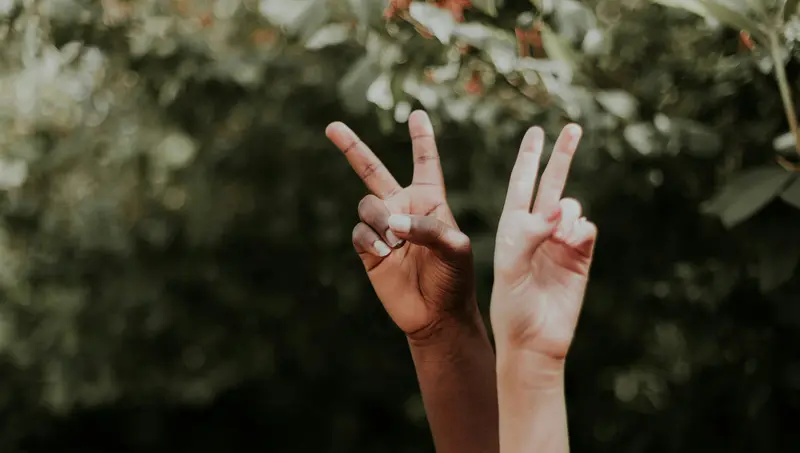 Two raised hands of different skin tones make peace signs against a blurred green, leafy background.