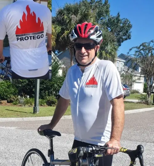 Un cycliste portant un casque rouge se tient debout à côté de son vélo de route, vêtu d'un maillot blanc PROTEGO, avec un empiècement représentant le grand logo PROTEGO en forme de flamme dans le dos.