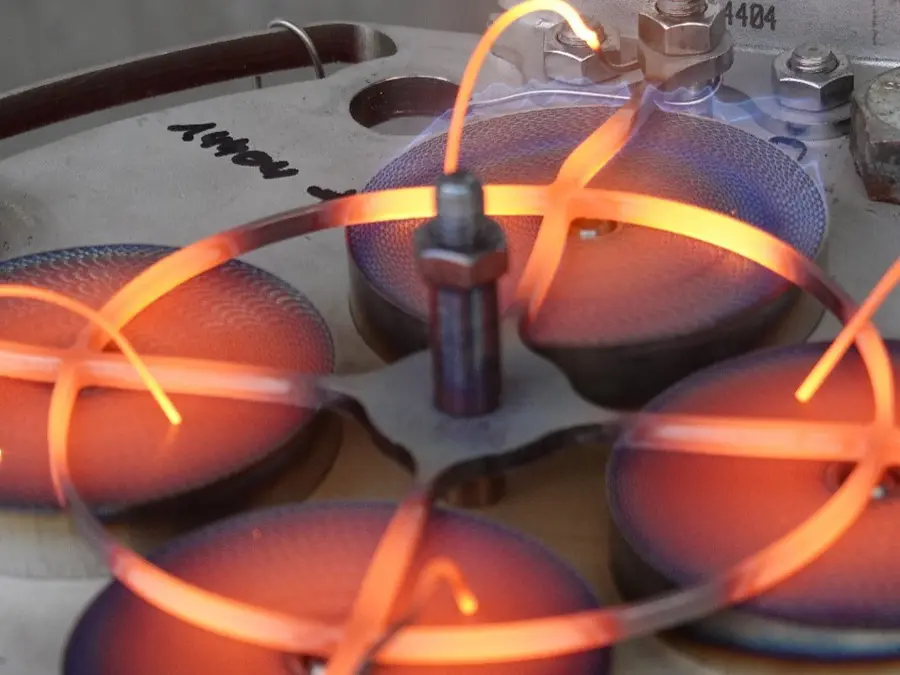 Close-up de um equipamento de teste de laboratório com fios aquecidos incandescentes e elementos metálicos circulares, mostrando uma configuração experimental ativa de alta temperatura.