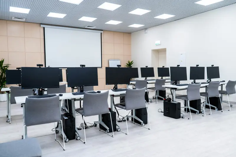 An empty computer lab classroom with rows of desks, desktop monitors, and a large projector screen at the front.