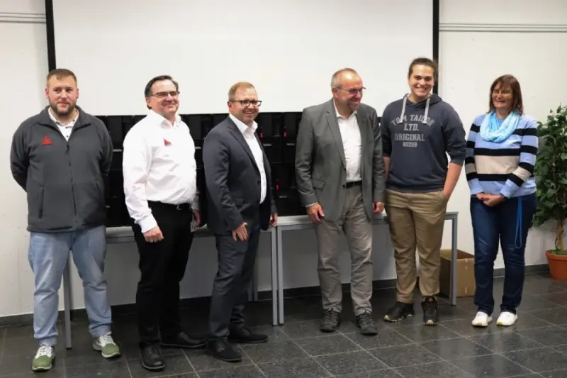 Sechs Personen stehen lächelnd für ein Gruppenfoto in einem Klassenzimmer, vor einem Tisch, auf dem schwarze Gerätekoffer und eine Projektionswand stehen.