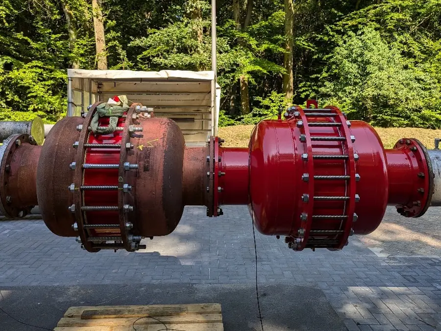 Comparação lado a lado de dois grandes equipamentos industriais com flange para tubulação ao ar livre, um fortemente corroído e outro recém-pintado de vermelho, montados entre tubos de aço contra um fundo arborizado.