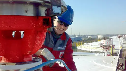 A worker in red coveralls walks through a large industrial plant filled with pipes, valves, and tall processing towers.