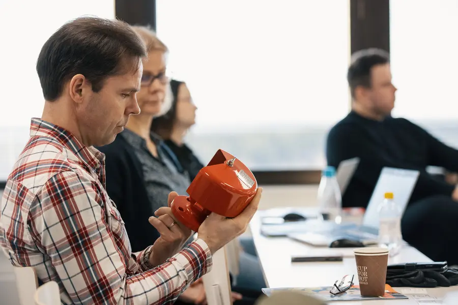 Ein Mann, der in einer schulähnlichen Umgebung sitzt, begutachtet eine rote Armatur, während andere Teilnehmer mit Notizbüchern und Laptops in der Nähe sitzen.