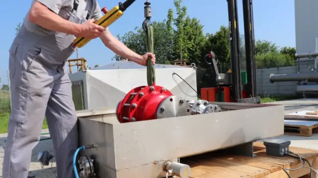 A technician lowers a large red industrial safety device into a rectangular outdoor test basin using a hoist, with cables and piping connected.