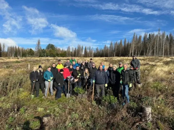 A large group of people wearing gloves pose with shovels in an open field, with sparse trees and a blue sky in the background.