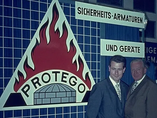 Two men in suits stand beside a large PROTEGO flame logo display and German signage reading “Sicherheits-Armaturen und Geräte” at an exhibition booth.