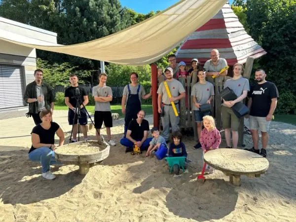 Un groupe d'adultes et d'enfants posent ensemble dans l'aire de jeux ensoleillée, sur le bac à sable, à l'ombre d'une grande voile d'ombrage, avec des équipements de jeux derrière eux.