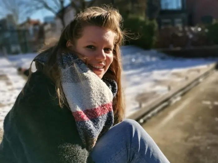 Une femme souriante, vêtue d'une épaisse écharpe rayée, est assise dehors par une journée d'hiver ensoleillée, avec de la neige et une allée qui se fondent dans l'arrière-plan.