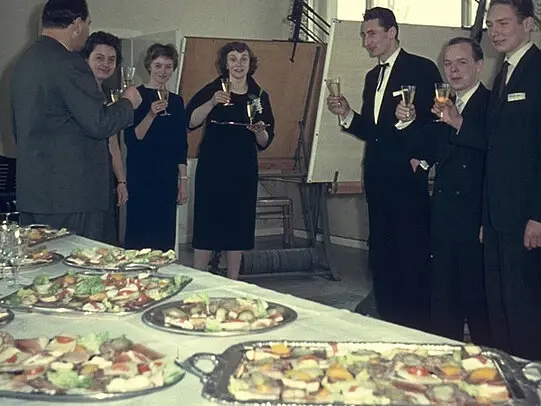 Vintage photo of formally dressed men and women raising glasses in a toast beside a table filled with assorted appetizers at a celebratory gathering.