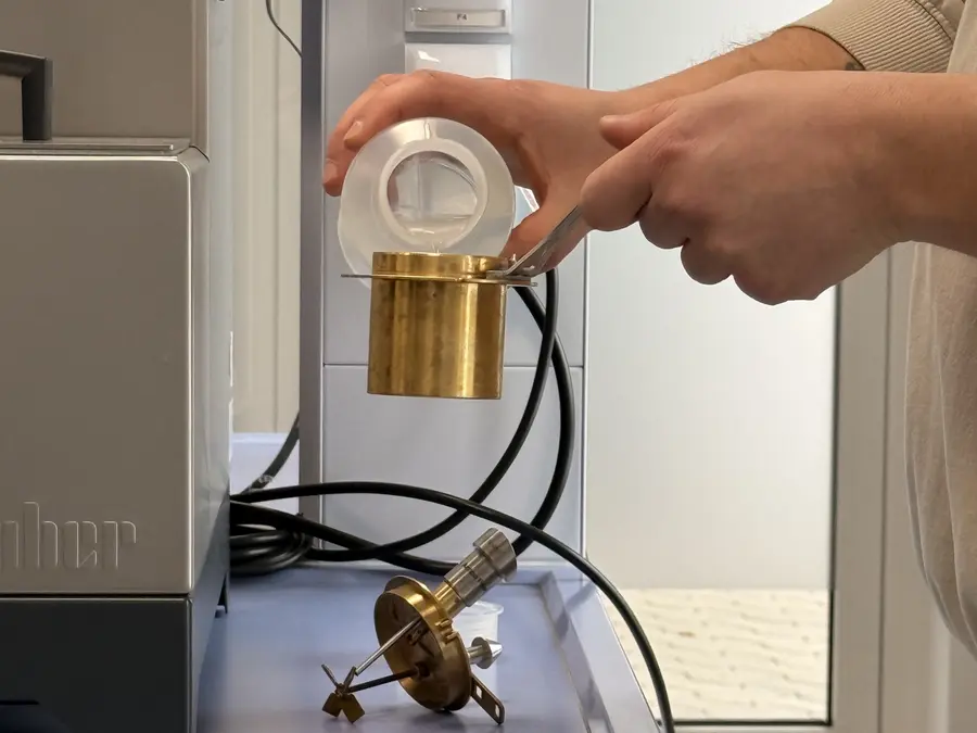 Eine Person hält einen kleinen Messingbecher mit einer Zange fest, während sie in einem Labor eine klare Flüssigkeit aus einem Plastikbehälter einfüllt.