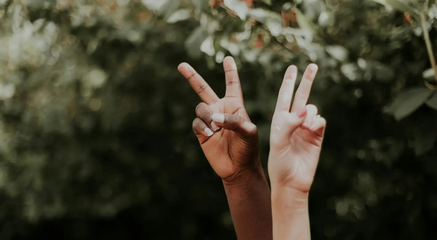 Deux mains levées, de teints différents, forment le signe de la paix sur un fond flou, vert et feuillu.