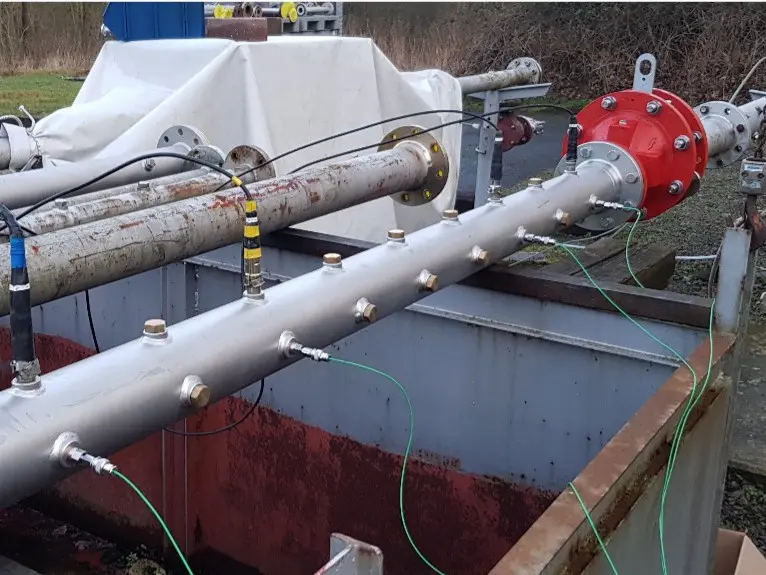 Configuração de teste industrial ao ar livre com um tubo horizontal sobre uma fossa de contenção aberta, apresentando um equipamento com flange vermelha e vários sensores conectados por cabos verdes.