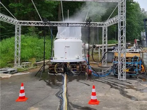 Instalação de testes industriais ao ar livre com um recipiente branco expelindo vapor sob uma estrutura de treliça metálica, cercada por mangueiras, equipamentos e cones de segurança.