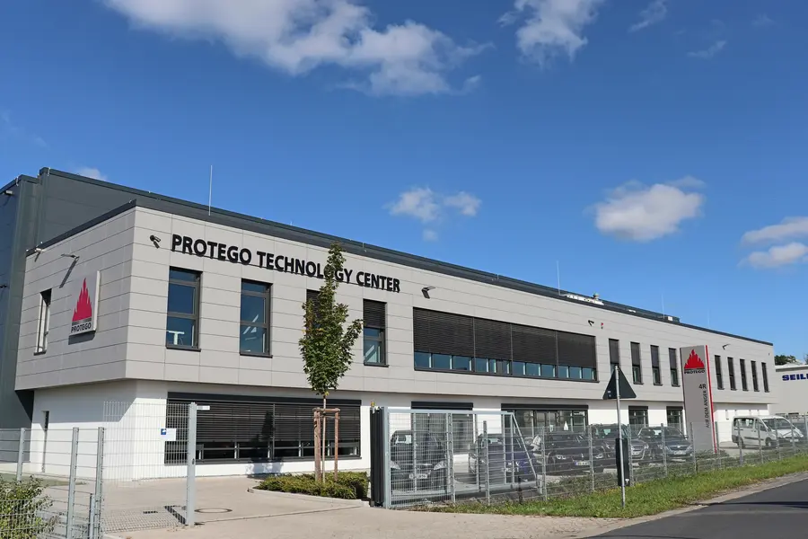 A modern office building labeled “PROTEGO TECHNOLOGY CENTER” under a blue sky, with a fenced parking area in front.