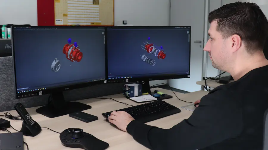A man works at a desk with dual monitors displaying 3D CAD models of a mechanical assembly, using a keyboard and control device.