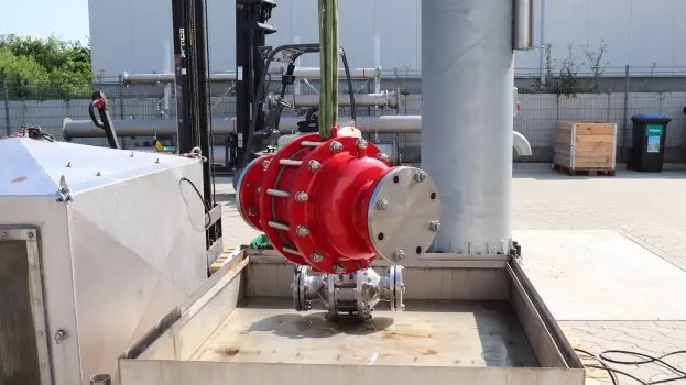 A large red industrial inline safety device is suspended by lifting straps above a metal test basin at an outdoor facility, with piping and equipment nearby.