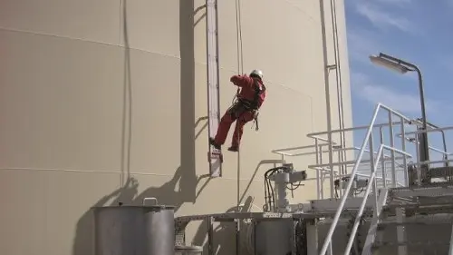 A rope-access worker in red gear climbs a ladder on the side of a large industrial tank, with metal platforms and piping below.