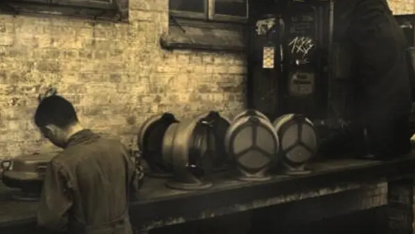 A vintage workshop scene shows workers at a long bench with several large cylindrical metal parts lined up against a brick wall.