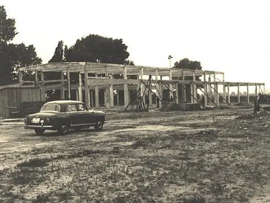 Vintage black-and-white photo of a car parked beside a partially constructed industrial building with exposed concrete framework.
