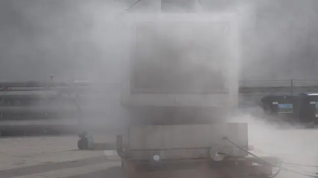 Outdoor industrial test setup with a large container largely obscured by a dense cloud of venting vapor or smoke during an active test.